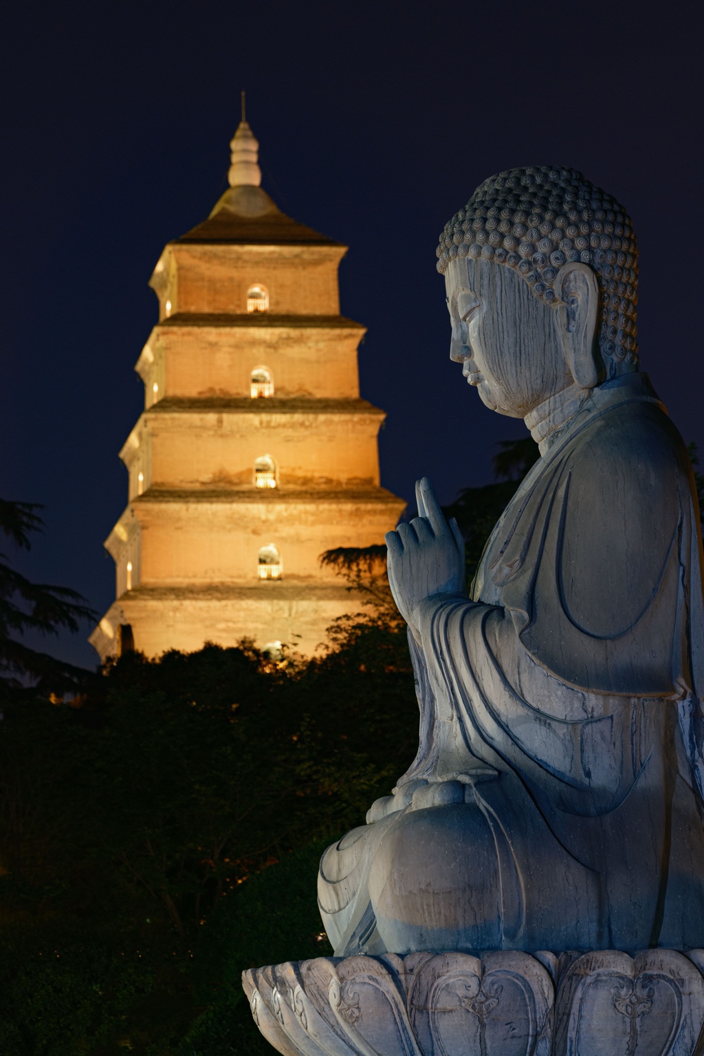A stone Buddha with the illuminated Big Wild Goose Pagoda behind it at night